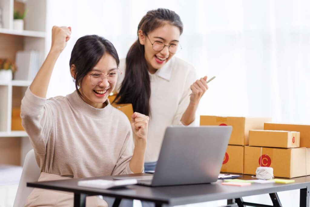 Two smiling Asian women in glasses, smiling in front of a laptop while surrounded by cardboard boxes.