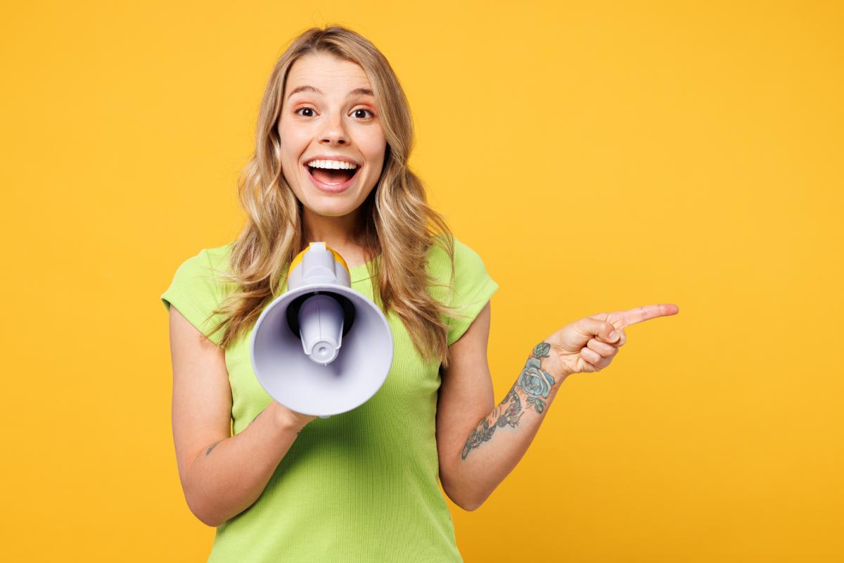 Young female direct sales representative stands smiling holding a megaphone wearing a green shirt set against a yellow background